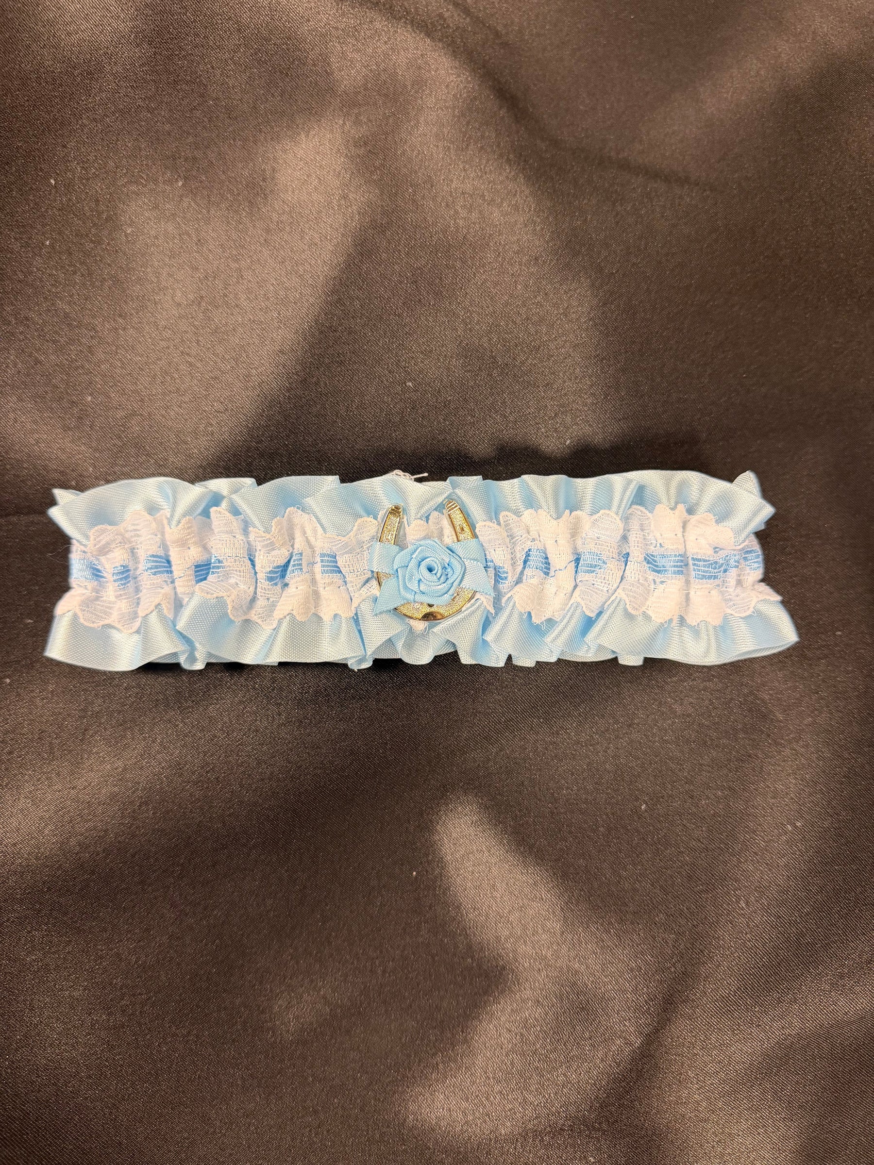 A light blue and white ruffled fabric garter with a central embroidered blue rose and gold accents, laid flat on a dark brown satin fabric background.