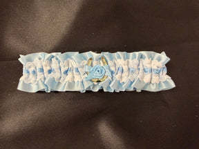 A light blue and white ruffled lace garter belt with a central blue fabric rose and gold-toned metal accents, displayed on a dark brown textured background.